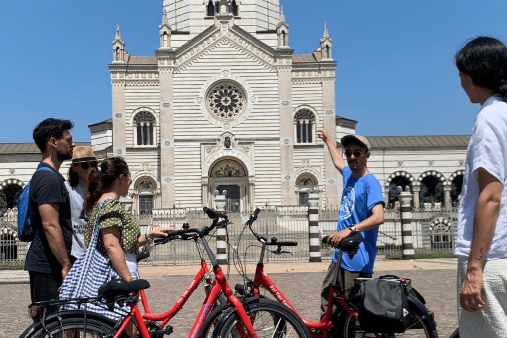 파란 하늘 아래 화려한 외관과 타워가 있는 유서 깊은 건물 앞에 빨간색 자전거를 탄 두 사람이 서 있습니다.