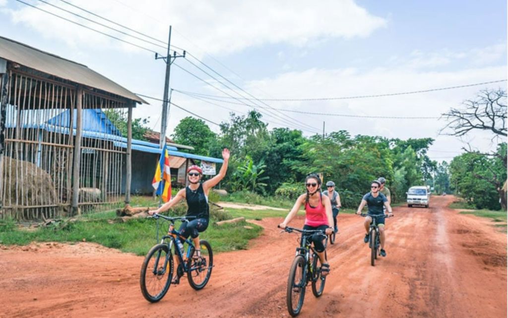 푸르름이 우거진 붉은 비포장 도로에서 자전거를 타는 4명의 승객이 차량을 배경으로 자전거를 탑니다. 한 명은 자전거를 타고, 다른 한 명은 페달을 밟으며 앞으로 나아갑니다.