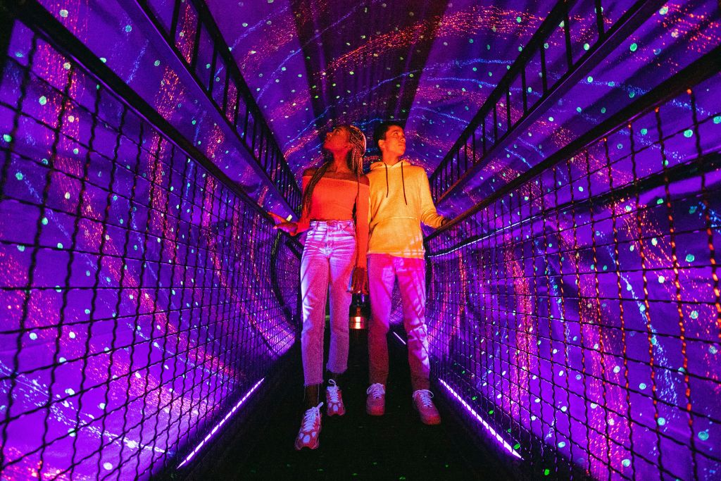 두 사람이 활기찬 보라색과 분홍색 조명이 있는 산책로 터널에 서서 천상의 우주 분위기를 연출합니다.