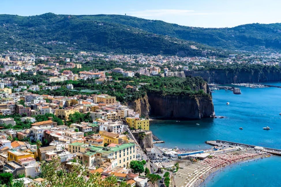 푸른 언덕을 배경으로 다채로운 건물, 모래 해변, 절벽, 깊고 푸른 바다가 펼쳐지는 해안 마을의 파노라마 전망.