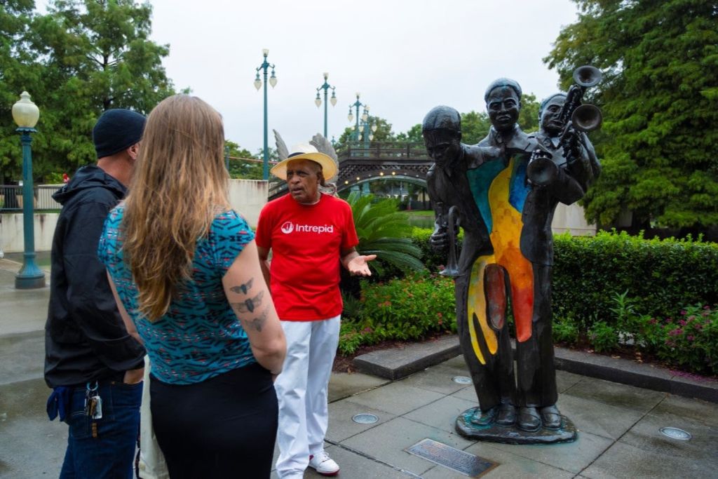 빨간색 인트레피드 셔츠를 입은 남성이 나무와 가로등이 있는 공원의 화려한 재즈 음악가 동상 근처에서 한 여성과 이야기를 나누고 있습니다.