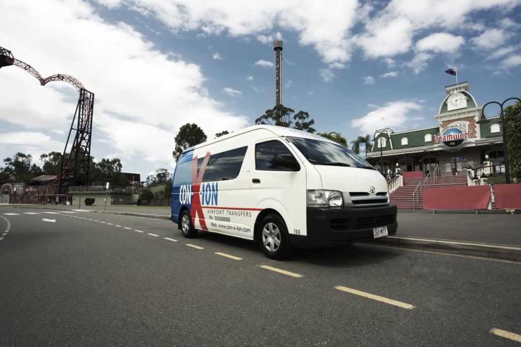 녹색 건물과 나무를 배경으로 거리에 주차된 흰색 Conn-X-ion 공항 환승 승합차.