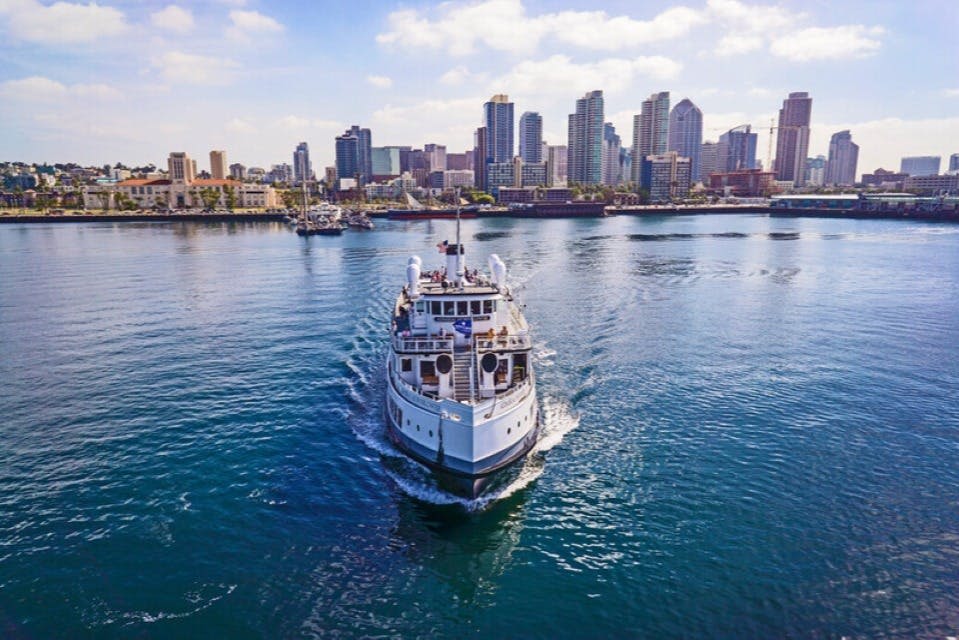 작은 유람선이 부분적으로 흐린 하늘 아래 도시 스카이라인을 배경으로 카메라를 향해 항해합니다.