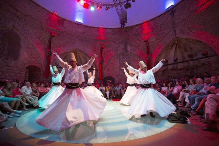 흰색 가운을 입은 댄서와 원뿔 모양의 모자가 원형의 조명으로 둘러싸인 방에서 관객들이 둘러앉아 공연을 펼칩니다.