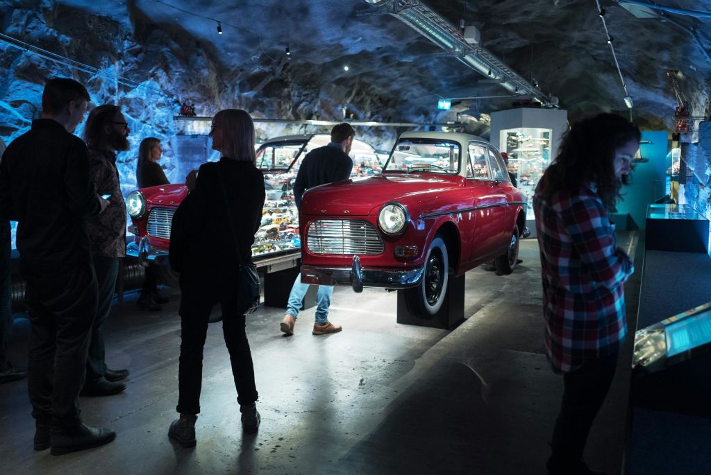 바위를 테마로 한 인테리어와 다양한 작은 전시물을 배경으로 실내에 전시된 빨간색 클래식 카를 관찰하는 사람들.