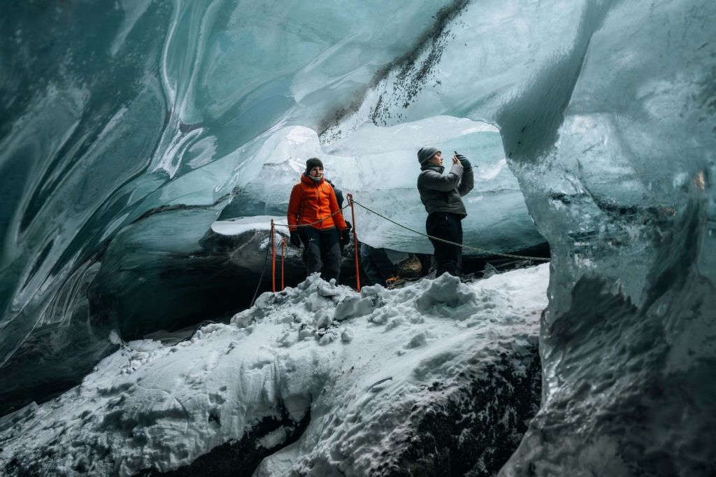 얼음 동굴 안에 서 있는 두 사람, 한 사람은 주황색 재킷을 입고 한 사람은 회색을 띠며 눈 덮인 얼음 벽으로 둘러싸여 있습니다.