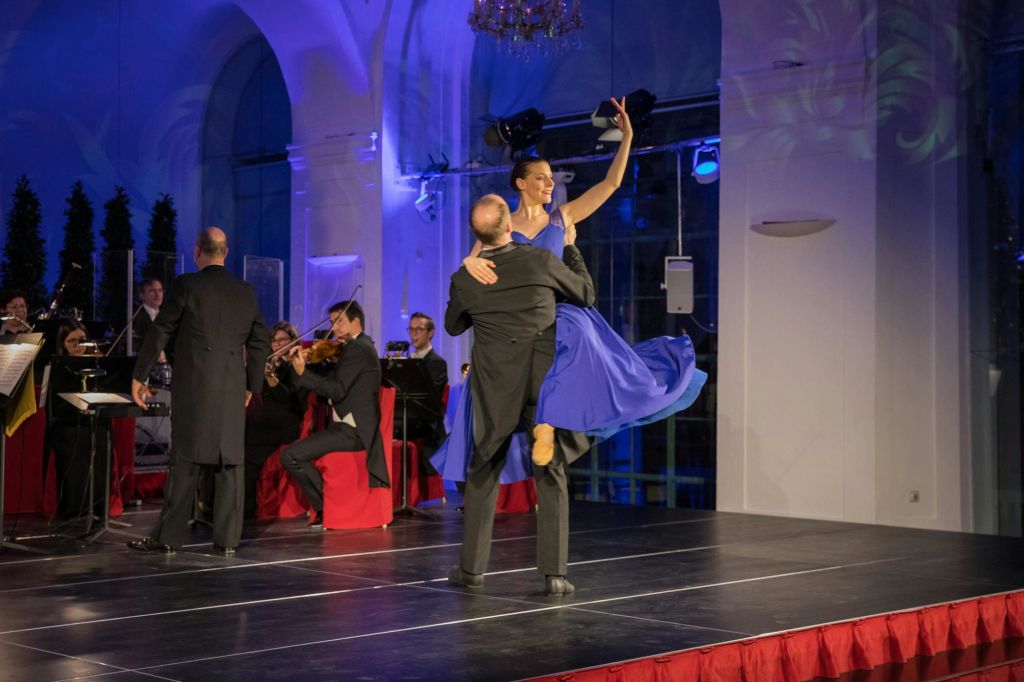 소규모 오케스트라가 무대를 배경으로 공연을 펼치는 가운데, 커플이 격식을 차려입고 무대에서 우아하게 춤을 추고 있습니다.