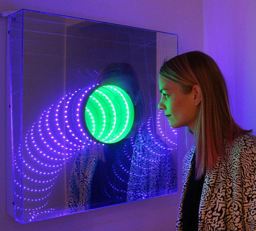 한 여성이 미러링된 설정에서 녹색과 보라색 LED 조명이 있는 다채로운 조명 예술 설치물을 자세히 들여다보고 있습니다.