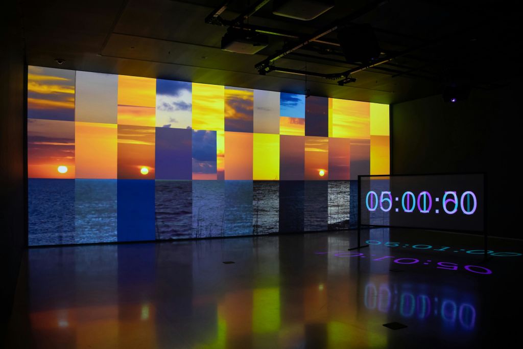 바다 위의 일몰을 세분화하여 투영한 현대 미술 작품으로 디지털 시계가 05:00 로 표시됩니다.