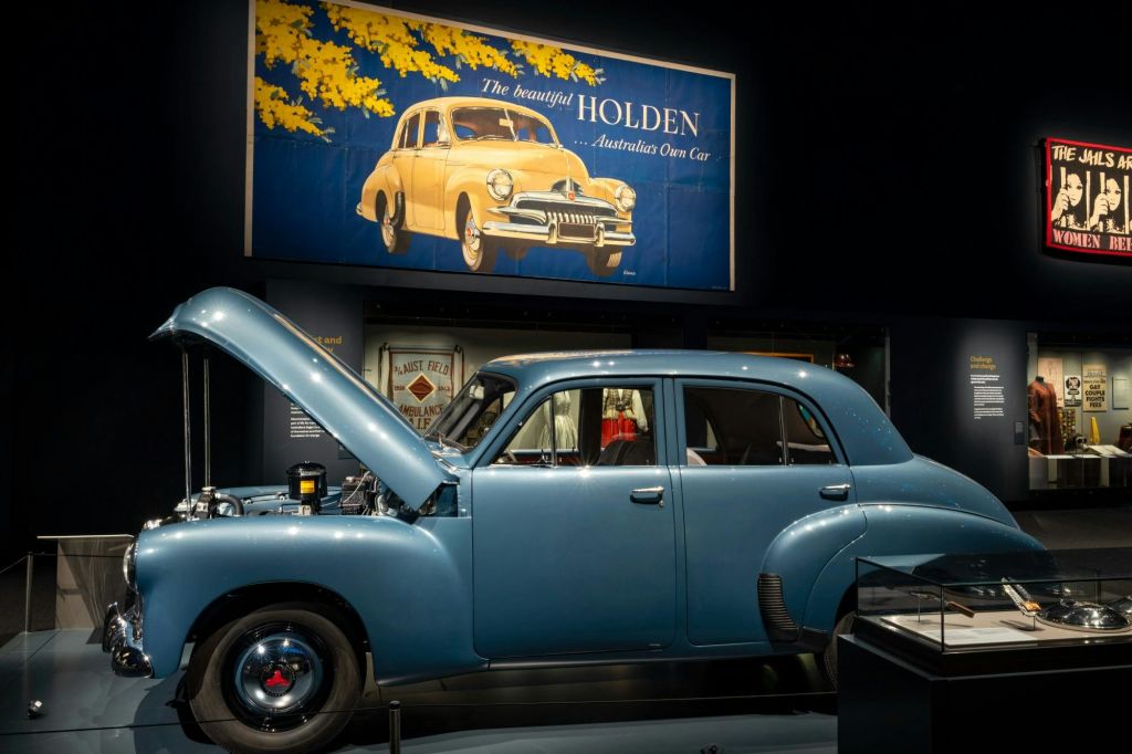 빈티지 블루 홀든 차량으로 후드가 열려 있으며, 박물관에 전시되어 있으며, 위에 레트로한 홀든 광고가 있습니다.