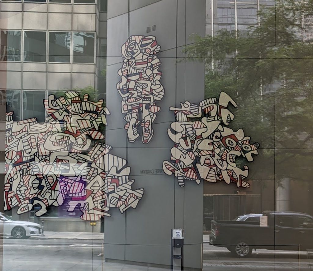 도시 거리와 건물이 배경으로 유리벽에 전시된 추상 미술 작품.