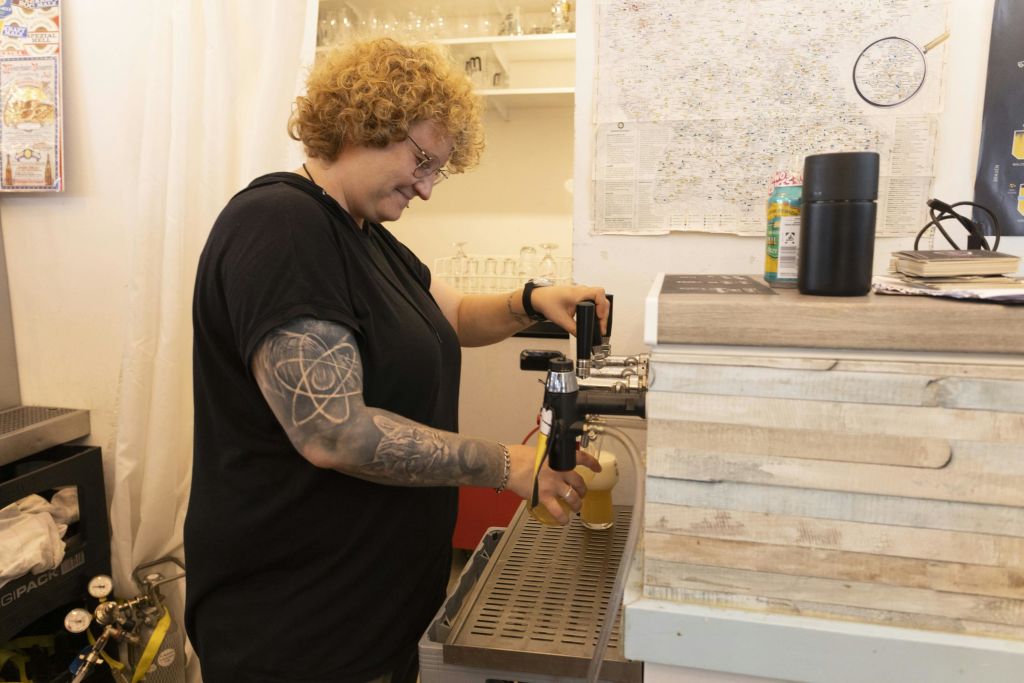 곱슬머리와 팔 문신을 한 사람이 바 뒤의 탭에서 음료를 쏟아내고 있습니다. 유리 제품과 지도가 있는 선반이 배경에 있습니다.