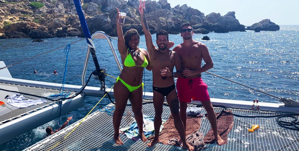 수영복을 입은 세 사람이 바닷가 보트 갑판에 서서 웃으며 바위 절벽을 배경으로 음료를 들고 있습니다.