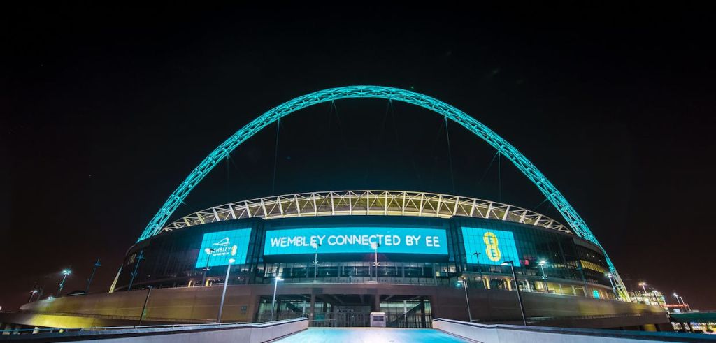 조명 아치와 "Wembley Connected by EE" 가 표시된 스크린이 있는 웸블리 스타디움의 야경.