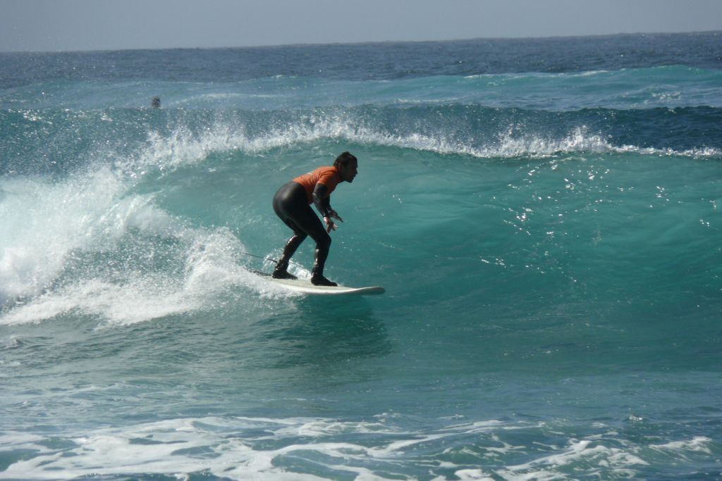 맑은 하늘 아래에서 웻수트와 오렌지 셔츠를 입고 커다랗고 푸른 물결을 타고 서핑하는 사람.