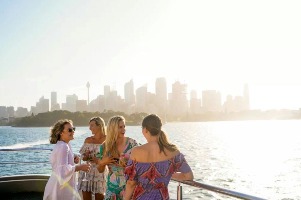 밝은 하늘 아래에서 도시의 스카이라인과 물을 배경으로 보트에서 사교하는 네 명의 여성.
