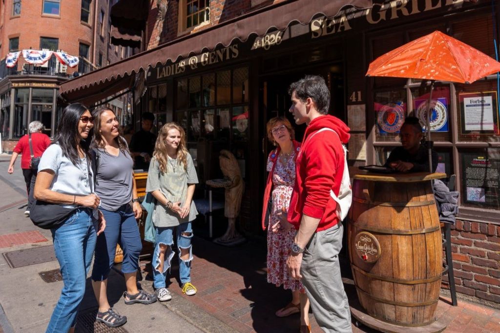 한 무리의 사람들이 "Ladies & Gents" 라고 적힌 천막이 있는 상점 앞 햇살 가득한 거리에 서서 이야기하고 있습니다.