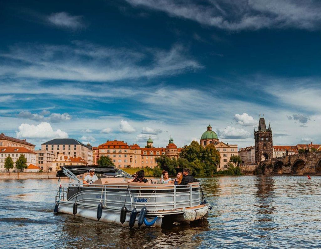 푸르고 흐린 하늘 아래 역사적인 건물과 탑을 배경으로 사람들이 탑승한 작은 보트가 강에 떠 있습니다.