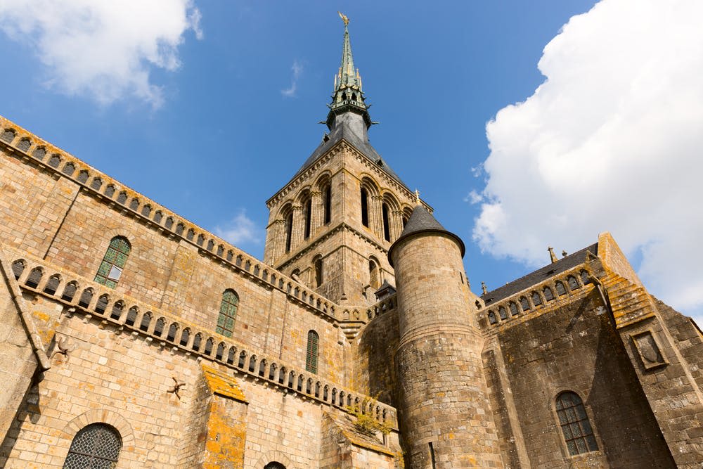 흰색 구름이 흩날리는 밝고 푸른 하늘 아래 높고 화려한 탑, 아치형 창문, 포탑이 있는 석조 건물.