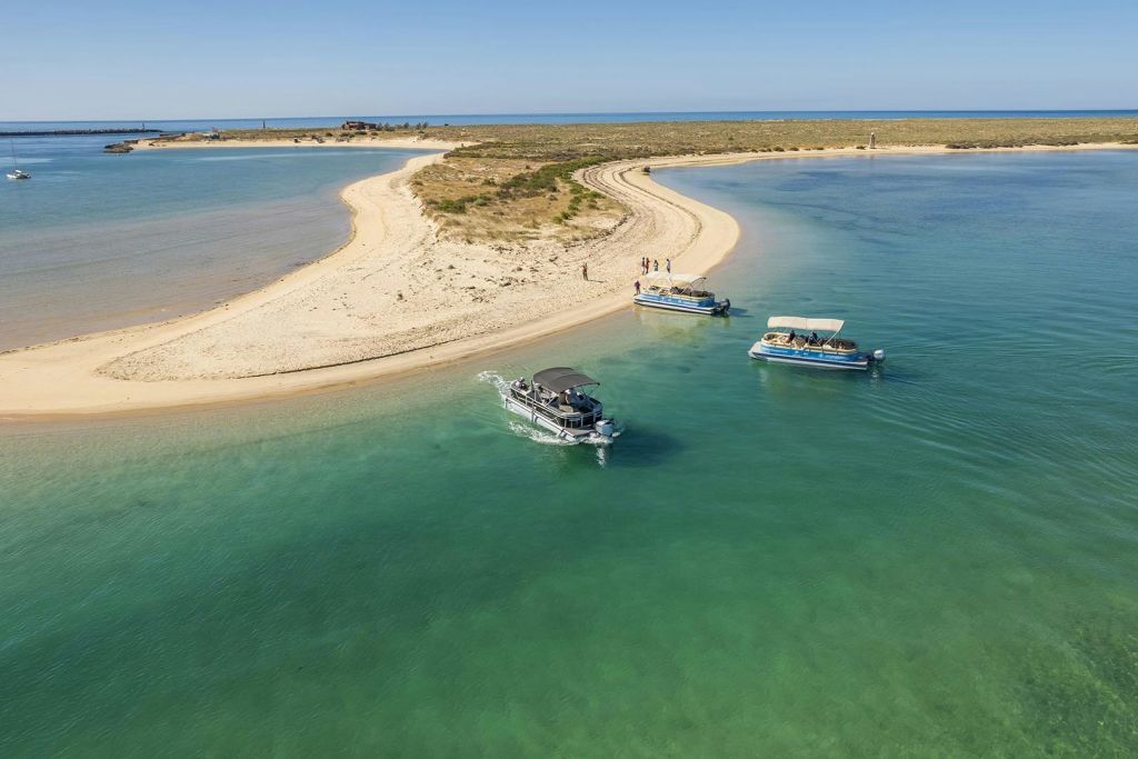 푸른 물이 흐르는 모래섬 근처의 프로모사마르 보트, 화창한 푸른 하늘 아래 해변을 거닐고 있는 방문객들.