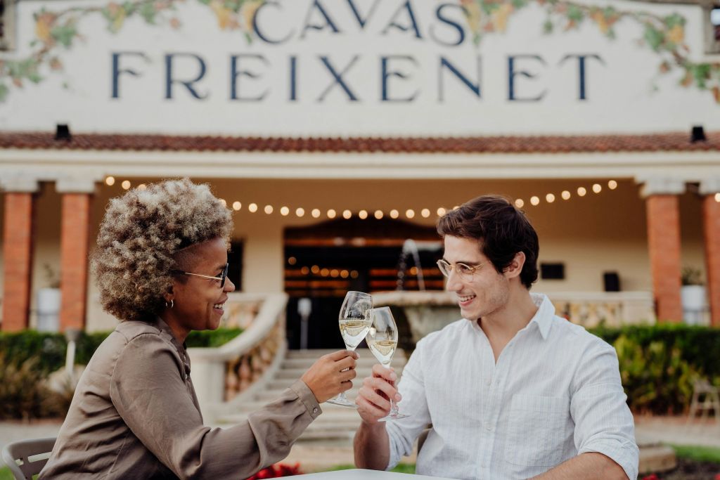 흰색 셔츠를 입은 남성이 "Cavas Freixenet" 간판이 있는 건물 앞에서 화이트 와인 한 잔을 들고 프레임에서 벗어난 사람과 건배합니다.