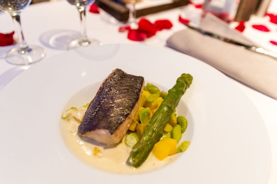 아스파라거스와 야채를 곁들인 생선 한 접시를 장미 꽃잎과 유리 제품을 배경으로 하얀 식탁보에 담았습니다.