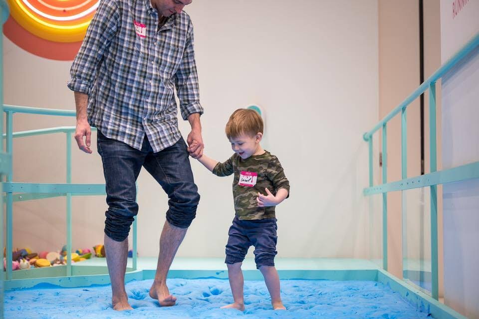 체크무늬 셔츠를 입은 성인과 카모 셔츠를 입은 어린이가 손을 잡고 파란색 패딩 바닥에서 맨발로 걷고 있습니다. 둘 다 이름 태그를 착용합니다.