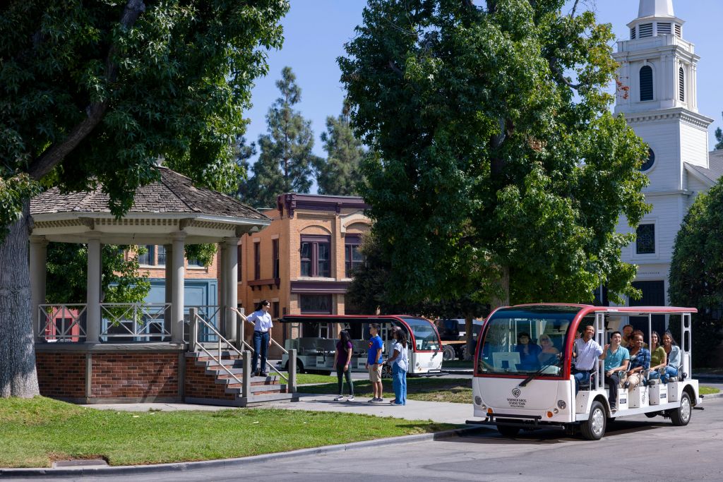 나무, 정자, 벽돌 건물을 배경으로 한 공원에서 작은 흰색 트램을 타는 사람들.