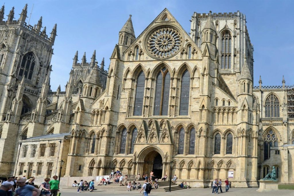 장미 창문, 아치형 출입구, 첨탑이 있는 화려하고 커다란 대성당 정면. 사람들이 바위 계단에 앉아 걸어가고 있습니다.