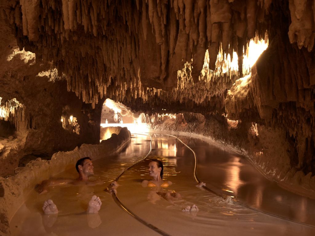 천장에 종유석이 매달려 있는 천연 지하 동굴 수영장에서 두 사람이 미소를 짓고 휴식을 취하고 있습니다.