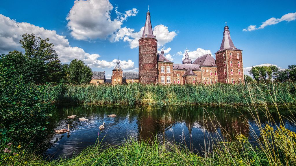 백조가 있는 반사 연못에 인접하여 녹지로 둘러싸인 포탑이 있는 유서 깊은 벽돌 성입니다. 구름이 있는 밝고 푸른 하늘.
