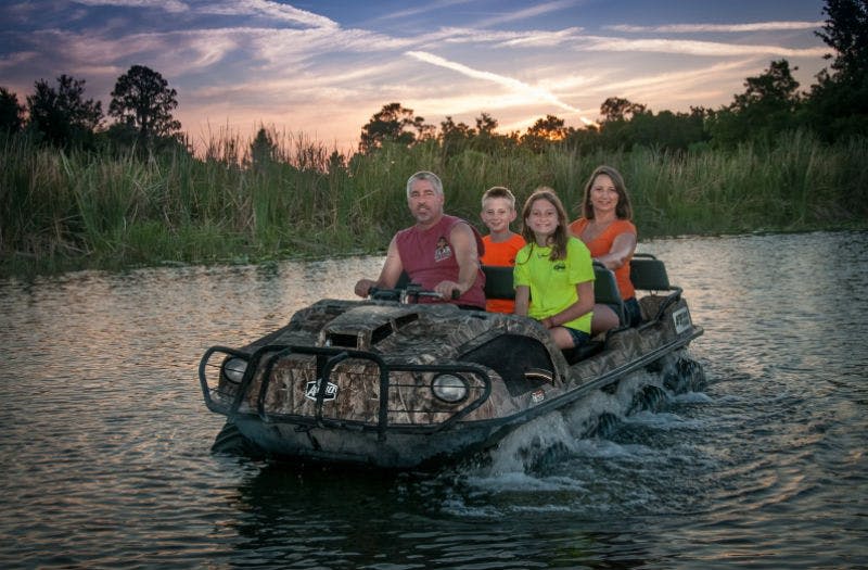 해 질 녘, 갈대와 나무를 배경으로 물 위에서 수륙양용 차량을 타고 있는 4인 가족의 모습입니다.