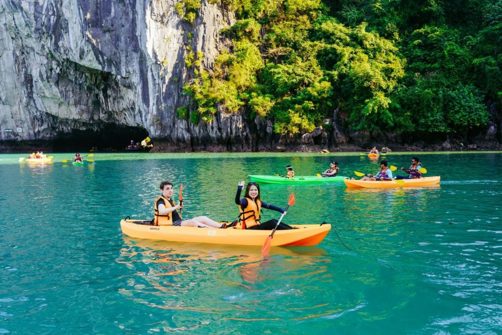 바위 절벽과 녹지 근처의 청록색 바다에서 노란색 카약을 탄 두 사람이 다른 카약 타는 사람들을 배경으로 노를 젓고 있습니다.