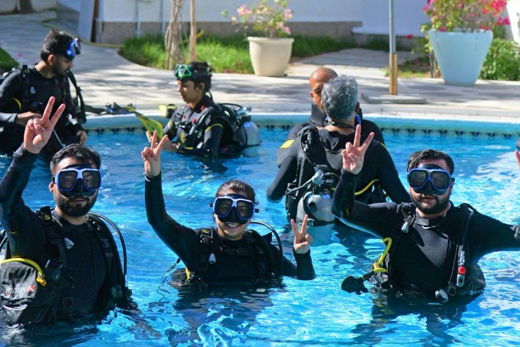 수영장에서 두 명의 스쿠버 다이버가 풀 장비를 착용하고 미소 지으며 평화 표지판을 만들고 다른 사람들은 백그라운드에서 연습합니다.