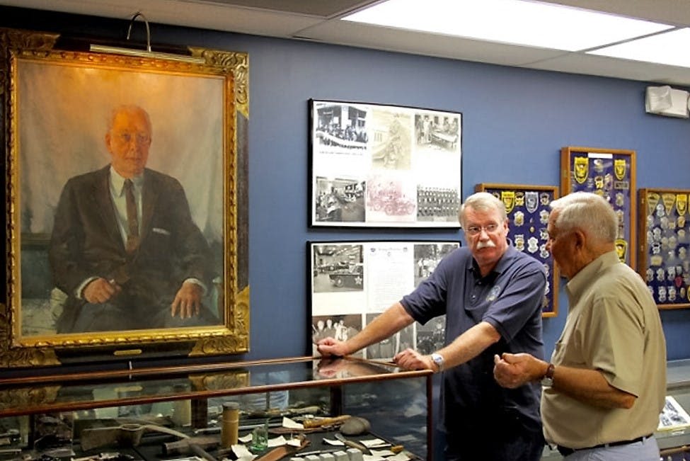 역사적 유물이 전시된 유리 진열장 옆에 한 남성이 커다란 초상화와 액자에 담긴 역사 사진 앞에 서 있습니다.