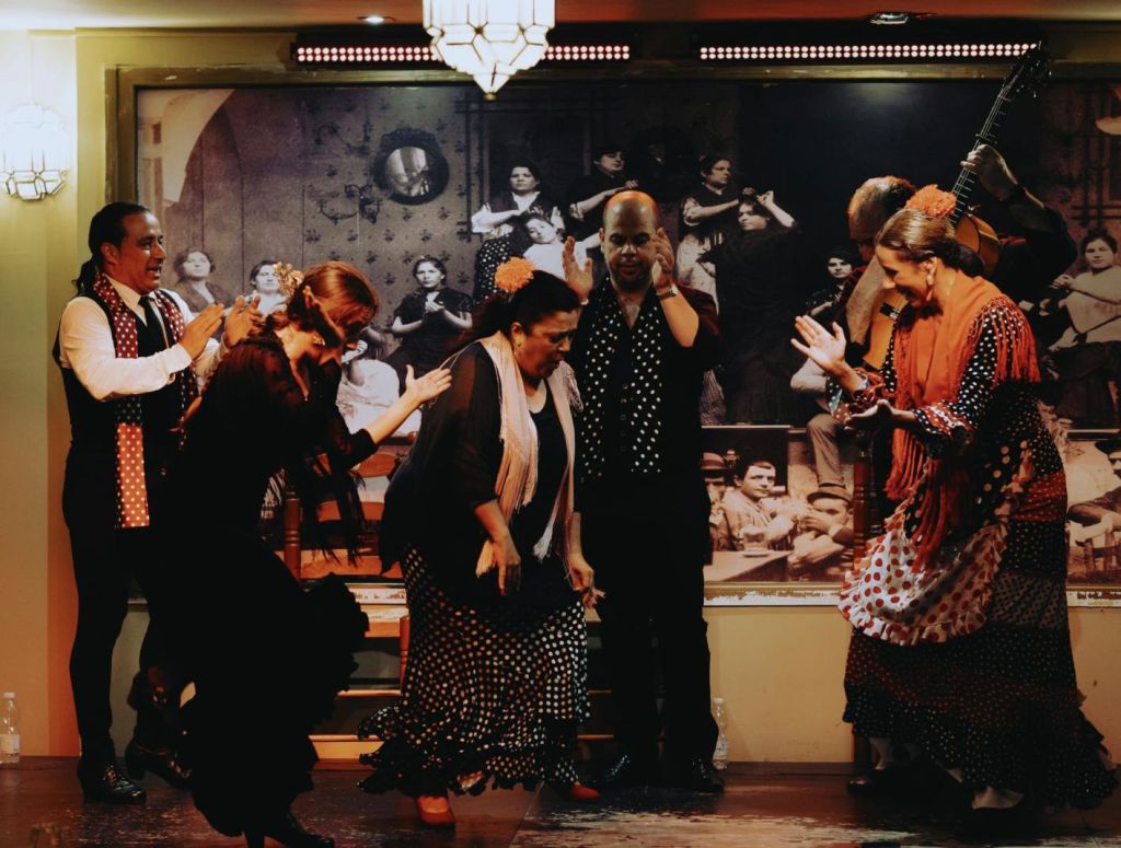 무대에서 전통 복장을 하고 열정적으로 춤을 추는 네 명의 사람들이 배경 사진에 관객과 함께합니다.