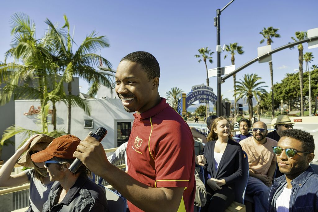 마이크를 들고 웃는 투어 가이드가 화창한 날씨에 해안가 근처에서 오픈탑 버스 투어를 진행합니다.