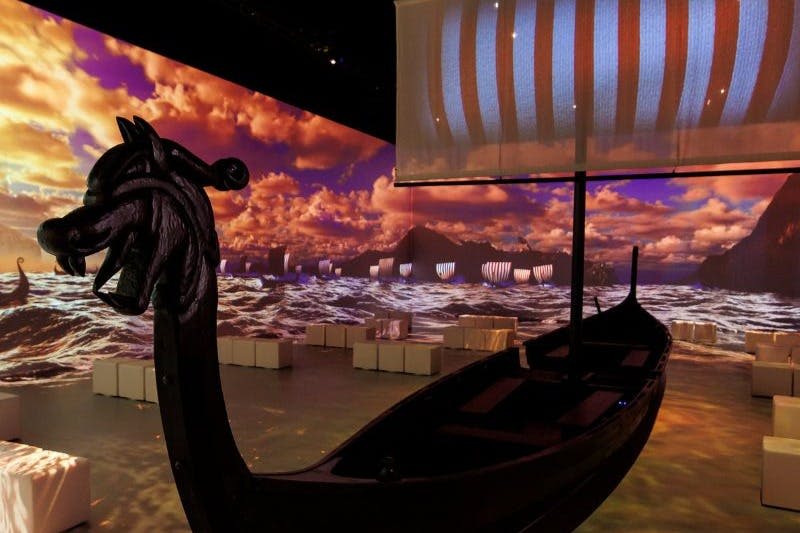 드라마틱한 하늘 아래 바다 풍경과 산을 배경으로 실내에 줄무늬 돛이 달린 바이킹 선박 복제품.