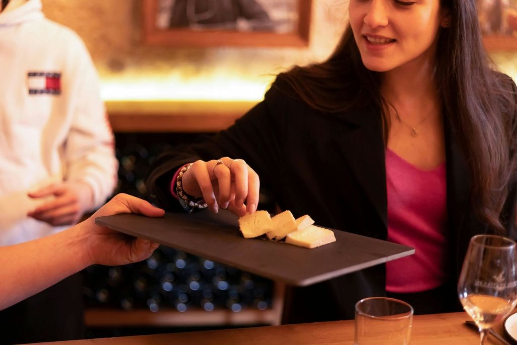 검은색 블레이저를 입은 여성이 치즈 조각이 담긴 슬레이트를 들고 있습니다. 나무 테이블에 와인잔과 접시가 보입니다.