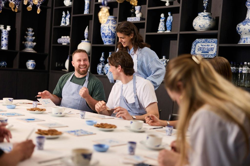 앞치마를 두르고 미소를 지으며 테이블에 도자기 그림을 그리는 사람들. 배경의 선반에 파란색과 흰색 도자기가 진열되어 있습니다.