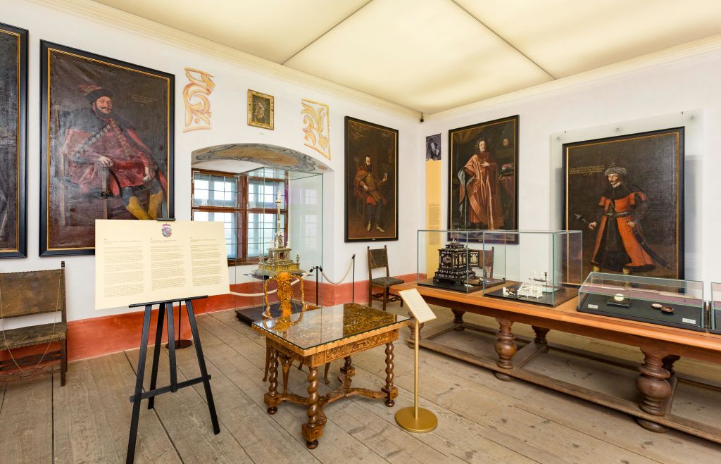액자에 담긴 초상화, 복잡한 모형이 들어 있는 전시 케이스, 화려한 테이블, 안내판이 있는 박물관 방.