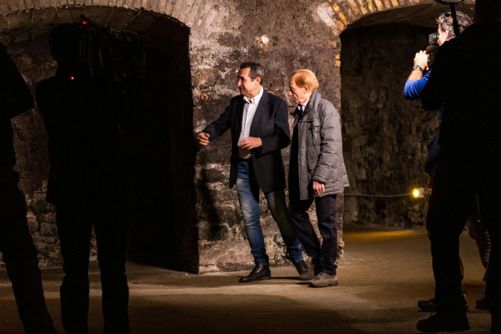 아치형 천장이 있는 어두운 석조 복도를 걷고 있는 두 남성. 한 남성은 블레이저와 청바지를 입고, 다른 한 남성은 회색 코트를 입고 있습니다.