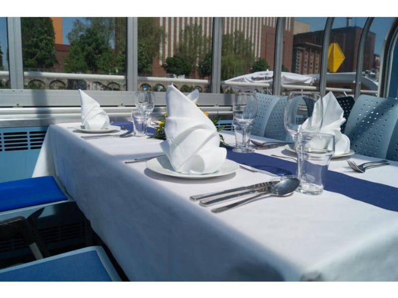 흰색 천, 접힌 냅킨, 와인잔, 식기류가 놓인 야외 식탁입니다. 배경에 도시적인 건물이 보입니다.