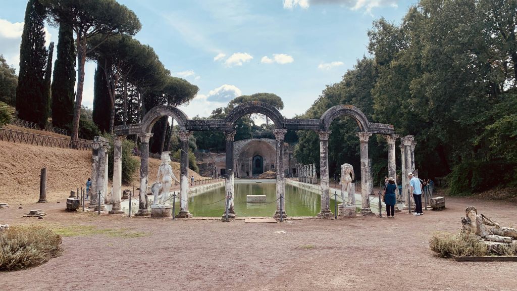 직사각형 수영장을 둘러싼 아치형 기둥이 있는 고대 유적지. 한 사람이 오른쪽 가장자리 근처에 서서 현장을 관찰합니다.