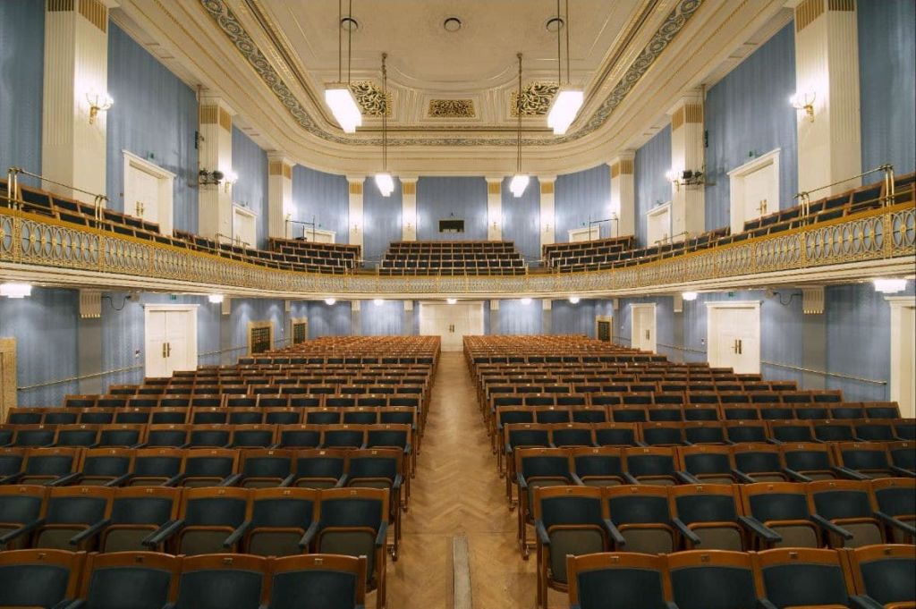 무대, 화려한 파란색 벽, 추가 좌석이 있는 발코니를 마주보고 있는 나무 좌석이 줄지어 있는 빈 강당.