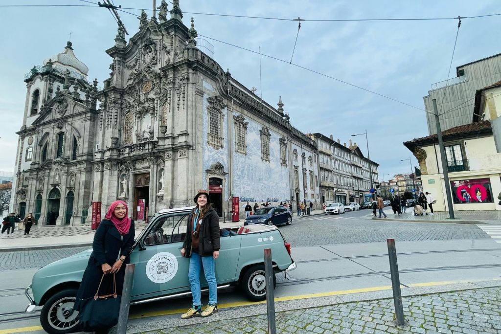 섬세한 건축미가 돋보이는 화려한 교회 건물 앞에 빈티지한 파란색 자동차 옆에 서 있는 두 사람.