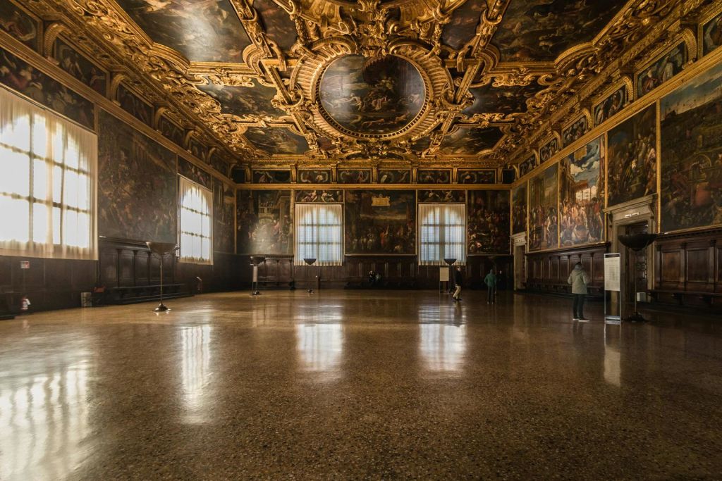 화려한 금과 페인트 천장, 커다란 창문, 벽 예술을 관찰하는 사람들이 있는 그랜드 홀. 바닥은 광택이 나고 반사됩니다.