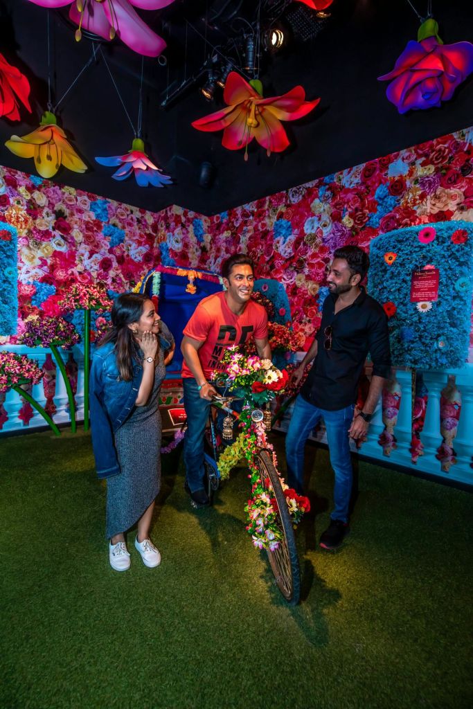 커다란 꽃이 걸려 있는 활기차고 꽃을 테마로 한 방에서 세 사람이 다채롭고 꽃으로 장식된 자전거와 상호작용합니다.