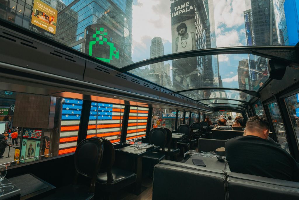 승객들은 창문과 천장을 통해 보이는 도시 건물과 광고판이 있는 유리 지붕이 있는 버스 안에 앉아 있습니다.