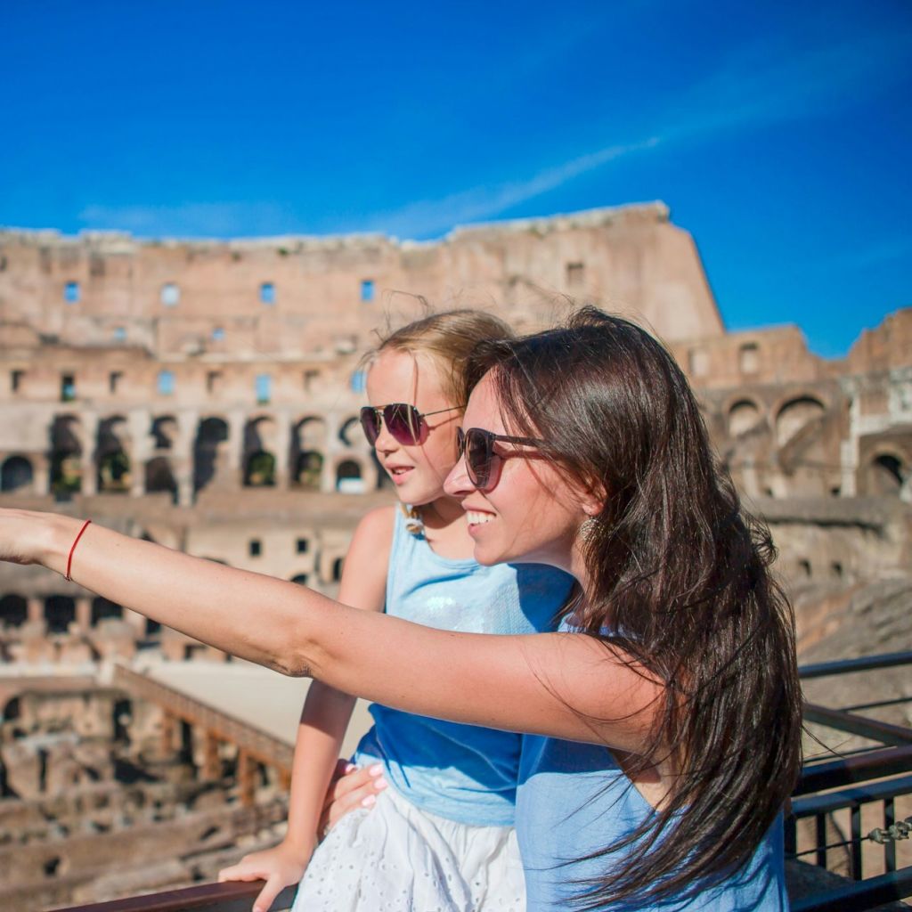 어른 한 명과 어린이 한 명, 선글라스를 착용한 두 사람이 맑고 푸른 하늘 아래 콜로세움 앞 난간 근처에 서 있습니다.